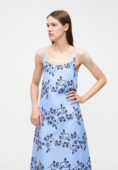 Γυναίκα με ίσια καστανά μαλλιά, που φοράει αμάνικο φωτεινό μπλε φόρεμα με σκούρες ανθρωπομορφικές κεντήματα, με το χέρι στη μέση, κοιτάζοντας προς το πλάι.