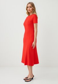 Een vrouw staat zijwaarts, gekleed in een strakke rode jurk met korte mouwen en een uitlopende zoom, gecombineerd met zwarte hoge hakken sandalen.