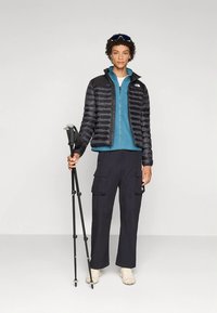 Črna puhaste zimska jakna preko modrega flisa z zadrgo, kombinirana s črnimi hlačami cargo. Stoji s pohodniškimi palicami, obut v lahke čevlje in z očali za zaščito.