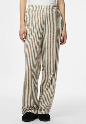 Kobieta ubrane w spodnie z wysokim stanem, luźne, z pionowymi beżowo-białymi paskami oraz czarne buty, stojąca na tle jednolitego białego tła.