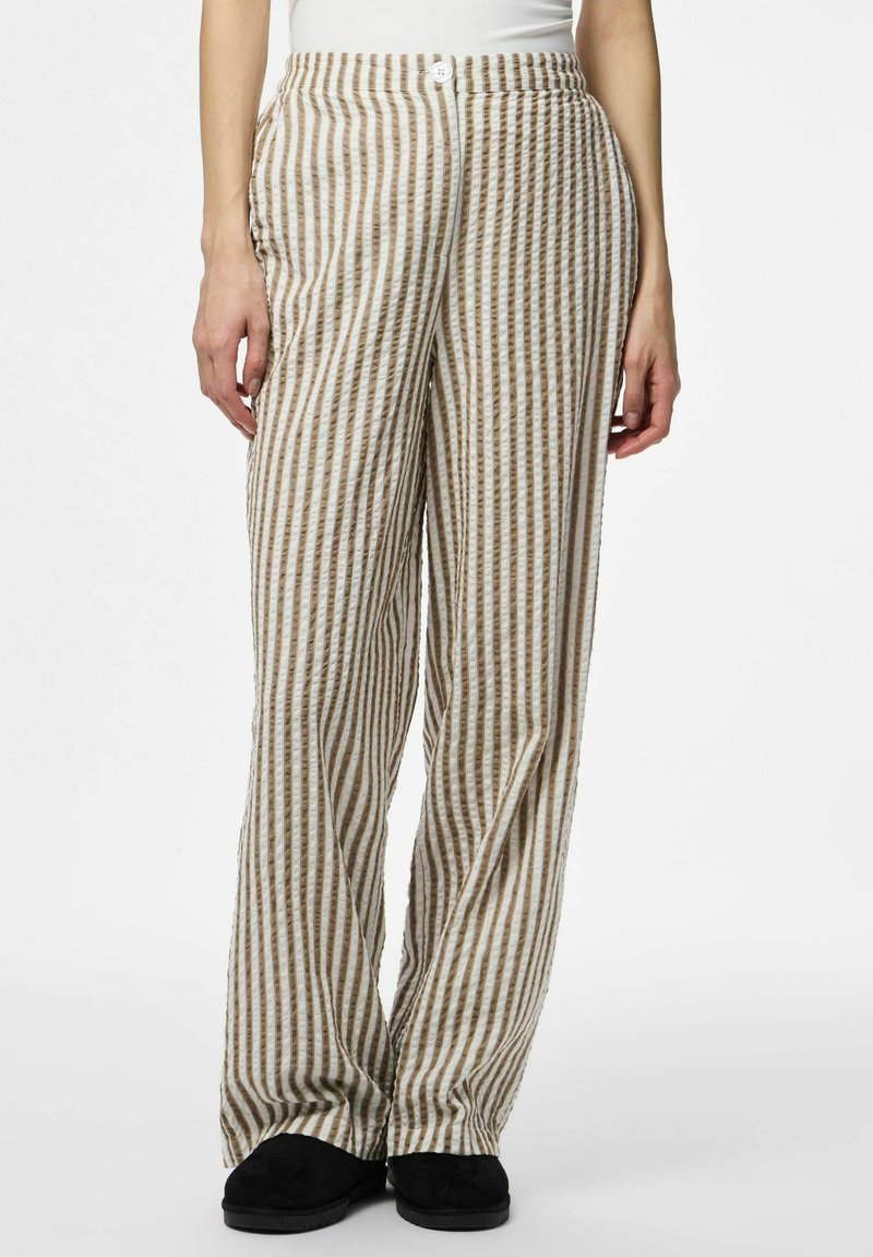 Femme portant un pantalon taille haute, ample, avec des rayures verticales beige et blanches, et des chaussures noires, debout devant un fond blanc uni.