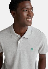 Šedé polo tričko z texturované látky, s klasickým límečkem, třemi knoflíky a zeleným logem na hrudi. Detailní pohled na výstřih.