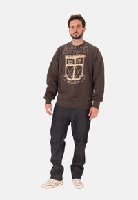 Felpa marrone con logo e motivo a stemma beige, abbinata a jeans blu scuro e sneakers bianche. Vestibilità comoda con colletto a coste.