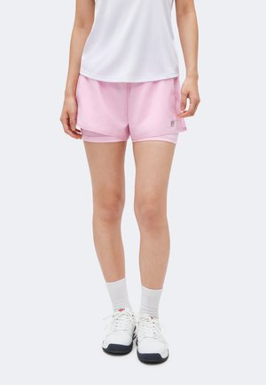 Person trägt hellrosa Sportshorts mit Netzeinsätzen an den Seiten, weißes Sporttop, weiße Socken und weiße Sneakers und steht vor einem einfarbigen Hintergrund.