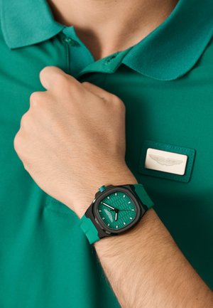 Zöld óra texturált számlappal, fekete tok, zöld szilikon szíj; mellé passzoló türkiz póló, címkével.