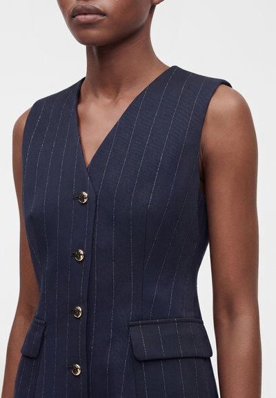 Femme portant un gilet sans manches bleu marine à fines rayures avec un col en V et des boutons dorés sur le devant.