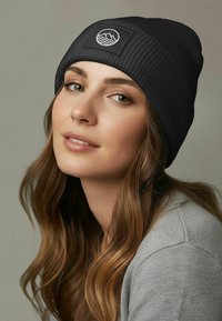 Young woman with long brown hair wears a black knit beanie with mountain and waves logo, a light gray top, against a plain neutral background.