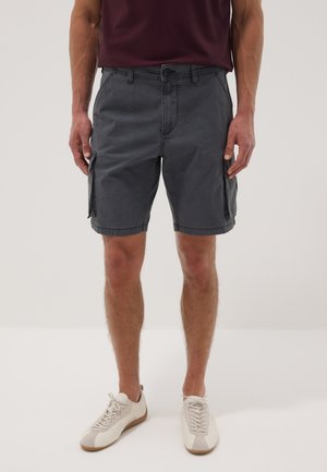 Uomo che indossa pantaloncini cargo grigio scuro, scarpe da ginnastica bianche e una camicia bordeaux, in piedi davanti a uno sfondo semplice.