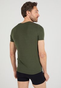T-shirt fitted di colore verde oliva con maniche corte, scollo rotondo e una texture di cotone morbido, indossata con slip neri, vista da dietro.