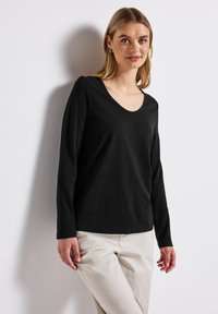 Schwarzes Langarmoberteil mit V-Ausschnitt, aus einem glatten Stoff gefertigt. Der Schnitt ist lässig und es wird mit hellen Hosen kombiniert.