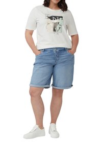 Frau in einem weißen T-Shirt mit Grafik, hellblauen, hochgekrempelten Jeansshorts und weißen Sneakers, die mit den Händen in den Taschen vor einem weißen Hintergrund steht.