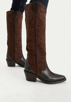 Brązowe kowbojki z miękkiej skóry i zamszu do połowy łydki, ozdobione haftowanymi wzorami i masywnym obcasem, noszone z ciemnoniebieskimi jeansami.