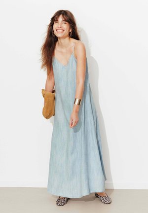 Femme souriante et penchée en avant, portant une robe maxi bleu clair sans manches, des ballerines imprimé léopard, un bracelet manchette doré, et tenant une pochette beige.