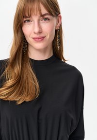 Czarna bluzka z wysokim kołnierzem, miękka w dotyku, z delikatnym układaniem. Długie kolczyki w stylu wiszących wisiorków z łańcuszkami i koralikami. Naturalne, rozpuszczone włosy.