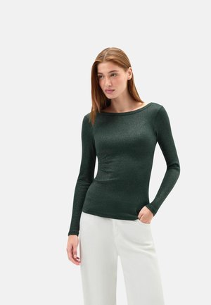 Donna con capelli lisci castano chiaro che indossa una maglia aderente verde scuro a maniche lunghe e pantaloni bianchi larghi su uno sfondo semplice.