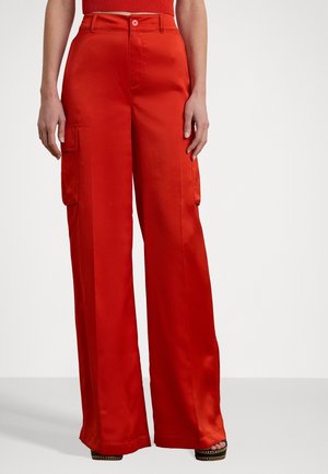 Person trägt hoch taillierte, weit geschnittene rote Hosen mit seitlichen Cargotaschen und ein passendes kurzes Oberteil, steht vor einem einfarbigen Hintergrund.