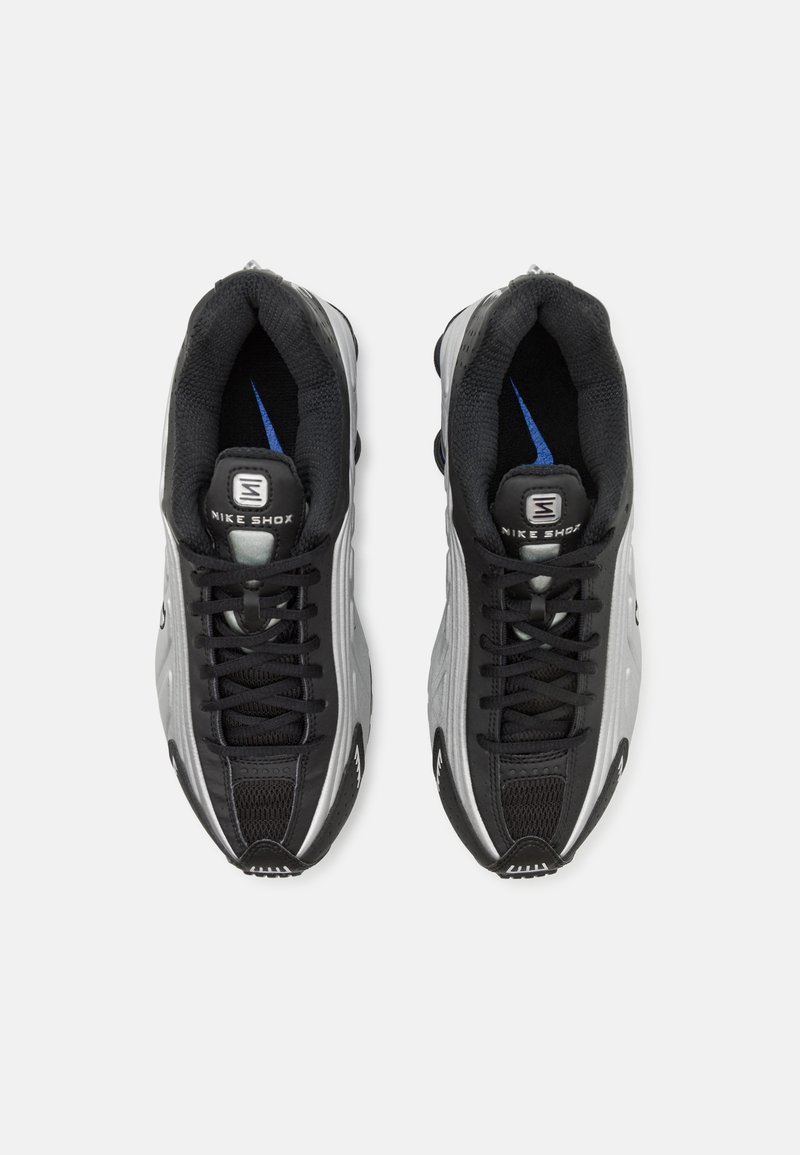 Baskets Nike Shox noir et argent avec un dessus texturé, un collier rembourré, des accents bleus et un système de laçage proéminent. La semelle présente un amorti distinct.