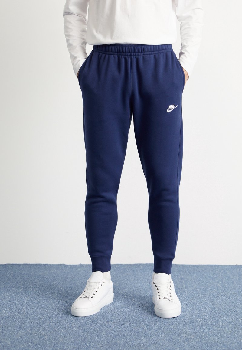 Námořnické modré Nike tepláky s elastickým pasem, slim fit a bílým logem. Spárovány s bílými teniskami na modrém texturovaném povrchu.