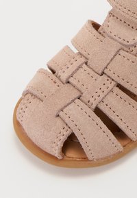 Ljusrosa mockasandal med ett vävt mönster, öppen tå och en platt brun sula. Sytt accentuerar den övergripande texturen.