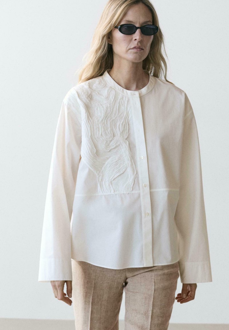 Femme aux longs cheveux blonds portant des lunettes de soleil noires, une chemise blanche ample à boutons avec une broderie texturée, et un pantalon beige clair.