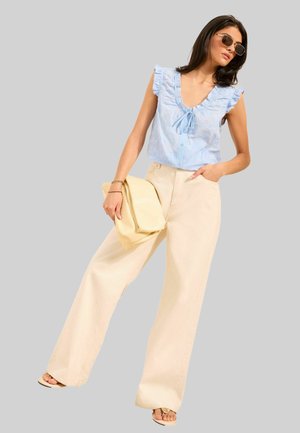 Kvinde iført lyseblå flæsetærklædet ærmeløs bluse, brede beige bukser, solbriller, holder en beige clutch, stående mod en ensfarvet baggrund.