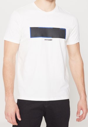 Mann trägt weißes Kurzarm-T-Shirt mit schwarzem rechteckigem Aufnäher, der blau umrandet ist, und dem Text "LIVE IN THE MOMENT" über schwarzer Hose.