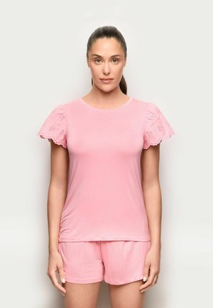 Frau mit dunklem Haar in einem Pferdeschwanz, die ein hellrosa Kurzarmshirt mit Lochspitzenärmeln und passende Shorts vor einem neutralen Hintergrund trägt.