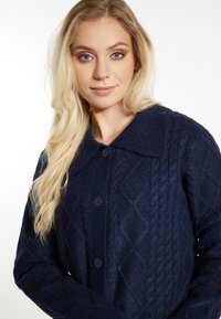 Marinblå stickad kofta med en diamantmönster, stor krage och knäppning. Mjuk textur med synliga kabelstickade detaljer.