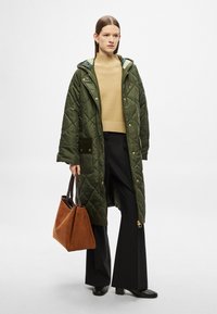 Zelený prošívaný dlouhý kabát s kapucí, béžový pletený svetr, černé kalhoty širokých nohavic a hnědá semišová taška přes rameno. Hladká textura, zlaté kovové detaily.
