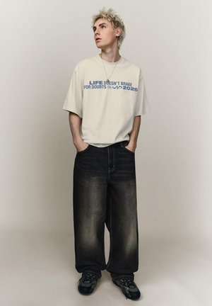 Junge Person mit kurzen blonden Haaren, die ein cremefarbenes T-Shirt mit blauem Text und lockere dunkle Jeans trägt, mit den Händen in den Taschen stehend und nach links blickend.