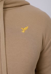 Maslinasta dukserica s rebrastom teksturom i prednjim zatvaračem, koja ima žuti izvezeni logo ptice na prsima.