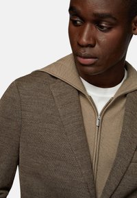 Brown fitted jacket with a zippered beige hoodie underneath. The fabric has a textured finish, displaying subtle patterns and stitching details.