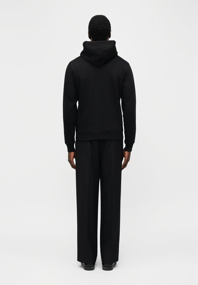 Άτομο που φοράει μαύρη μπλούζα με κουκούλα και φαρδιά μαύρη παντελόνια, στέκεται με την πλάτη προς τα έξω μπροστά σε ένα απλό φωτεινό φόντο.