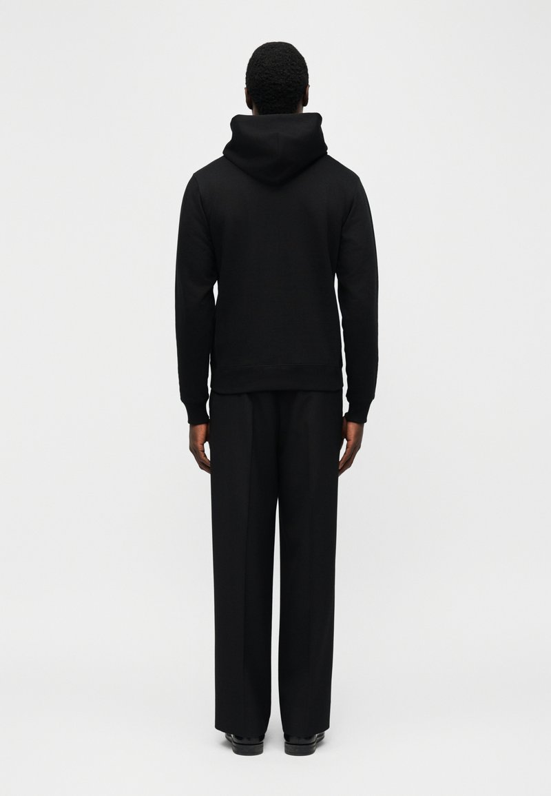 Person trägt schwarzen Hoodie und lockere schwarze Hose, steht mit dem Rücken zur Kamera vor einem einfachen hellen Hintergrund.