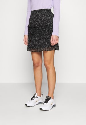 Černá sukně s volány a bílými puntíky, doplněná světlofialovým tričkem s dlouhým rukávem a bílými sportovními botami s černými tkaničkami a zlatými detaily.