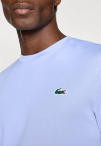 Μαυρου καφέ κοντομάνικο πουκάμισο με στρογγυλή λαιμόκοψη και μικρό πράσινο λογότυπο κροκόδειλου στην αριστερή πλευρά του στήθους. Λεία υφή.