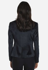 El blazer de terciopelo negro presenta un diseño ajustado, una textura suave y una abertura trasera sencilla, con puños abotonados y una silueta entallada.