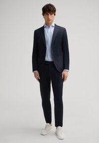 Granatowy garnitur z klapami w kształcie litery V, noszony na jasnoniebieskiej koszuli. Uzupełniony slim fit spodniami i białymi sneakersami dla casualowego wyglądu.