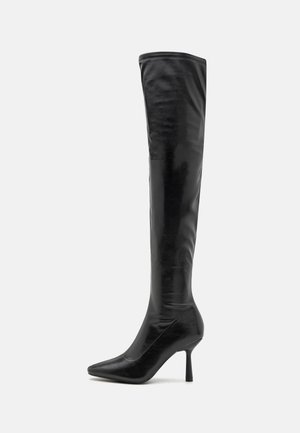 Botte en cuir noire montant jusqu’à la cuisse avec bout pointu et talon haut et fin sur fond uni gris clair.