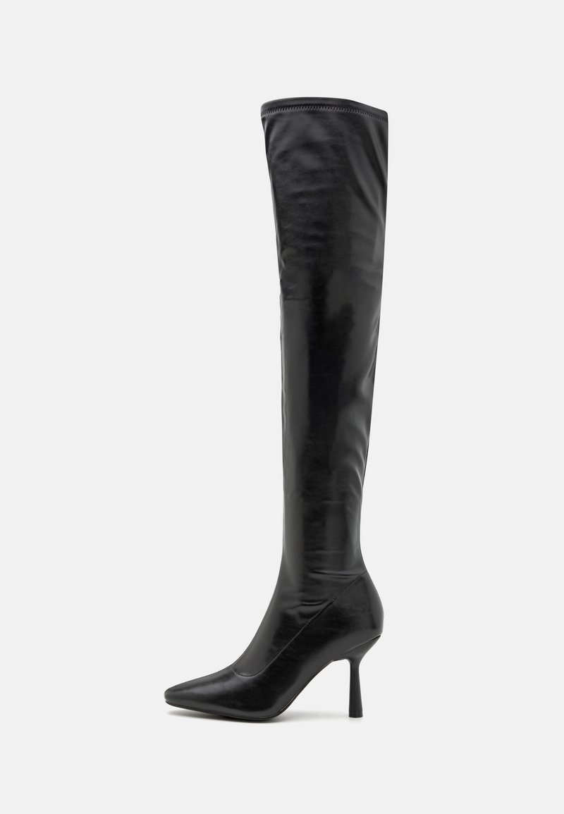 Botte en cuir noire montant jusqu’à la cuisse avec bout pointu et talon haut et fin sur fond uni gris clair.