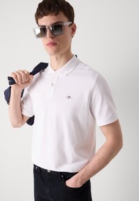 Hvit polo-skjorte med korte ermer, tre-knaps knappestolpe og logo-detalj. Modellen holder en marineblå jakke med den ene hånden, iført store solbriller.