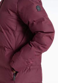 Burgunderfarbene Steppjacke mit einem auffälligen Badge auf der Schulter, strukturierter Oberfläche und einer Reißverschlusstasche an der Seite.