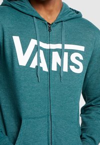 Felpa con zip color teal realizzata in tessuto morbido, caratterizzata da un logo "VANS" bianco sulla parte anteriore. Presenta un cappuccio con coulisse e tasche frontali.