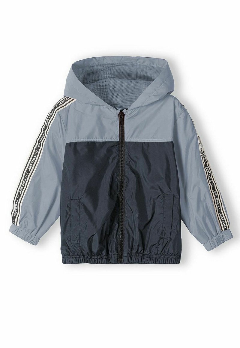 Veste coupe-vent bleue et marine avec capuche, fermeture éclair, poches latérales et accents rayés avec le texte "JUST CHILL" sur les manches.