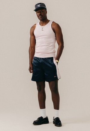 Homme portant un débardeur blanc, un short bleu marine et blanc, des chaussettes blanches, des chaussures noires, une montre en or, un collier et une casquette rayée bleu marine, debout devant un fond uni.