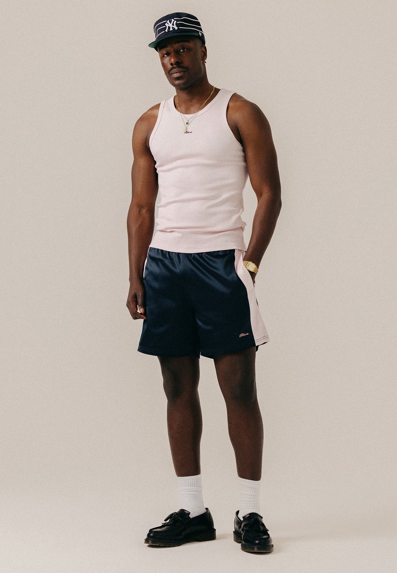 Homme portant un débardeur blanc, un short bleu marine et blanc, des chaussettes blanches, des chaussures noires, une montre en or, un collier et une casquette rayée bleu marine, debout devant un fond uni.
