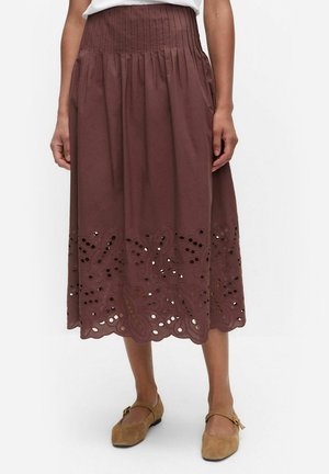 Bruine rok tot kniehoogte met geplooide tailleband en bloemmotieven uitgesneden langs de gegolfde zoom, gedragen met beige Mary Jane ballerina's.