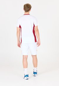 Hvid atletisk skjorte med røde sidelister, parret med hvide shorts med orange accenter. Inkluderer sokker og blå sportssko.