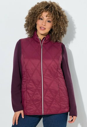 Vrouw met krullend haar draagt een doorgestikte bordeauxrode vest over een donkerpaars shirt met lange mouwen en blauwe spijkerbroek, staand tegen een witte achtergrond.