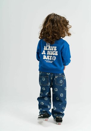 Dítě s kudrnatými vlasy má na sobě modrou mikinu s nápisem "Pěkný den" a vzorované džíny s usměvavými obličeji, stojí na bílém pozadí.